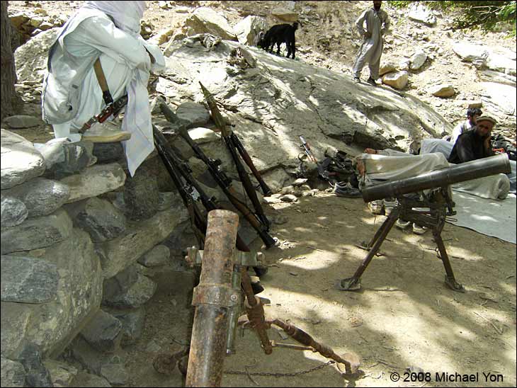 Foreground is a corroded mortar; leaning against the rocks is an old rifle and RPGs.  Background, the man appears to be sleeping with his head on French body armor (the high resolution images allow zooming in), and there appears to be a French helmet on the rocks next to the tennis shoes.  The AK-47 is affixed with the bayonet.  Did this bayonet kill some of the French? (At least one French soldier was killed by a blade.) Did this man get the gear because he killed the owner?  Did they strip the soldiers first so that the gear would not be bloodied?