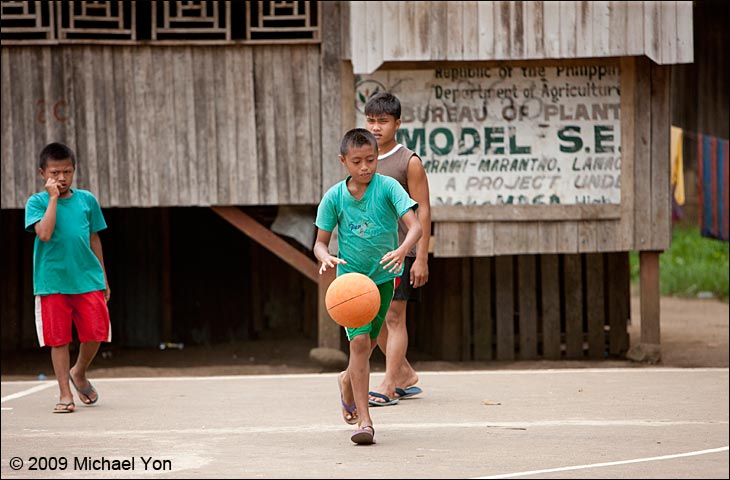 Enemy kids playing basketball in village by old sign.